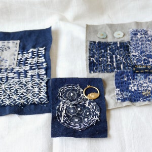 May include: Three blue fabric squares with white embroidery. The squares are stitched together with different patterns. One square has the text "Made in Japan" on it. The squares are on a white background.