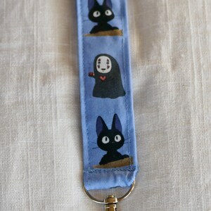 May include: A blue fabric keychain with a black cat, a white-faced character with a red heart, and another black cat. The keychain has a silver metal clasp.
