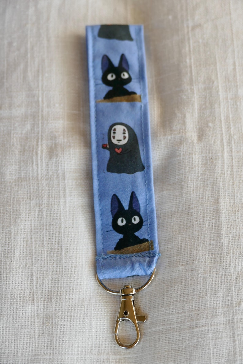 May include: A blue fabric keychain with a black cat, a white-faced character with a red heart, and another black cat. The keychain has a silver metal clasp.