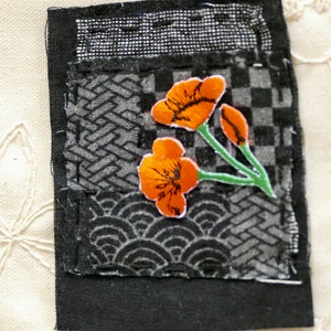 Peut inclure: Un carr&eacute; de tissu noir avec un motif patchwork pr&eacute;sentant diff&eacute;rents motifs. Une petite fleur orange brod&eacute;e avec des tiges vertes est centr&eacute;e sur le tissu.