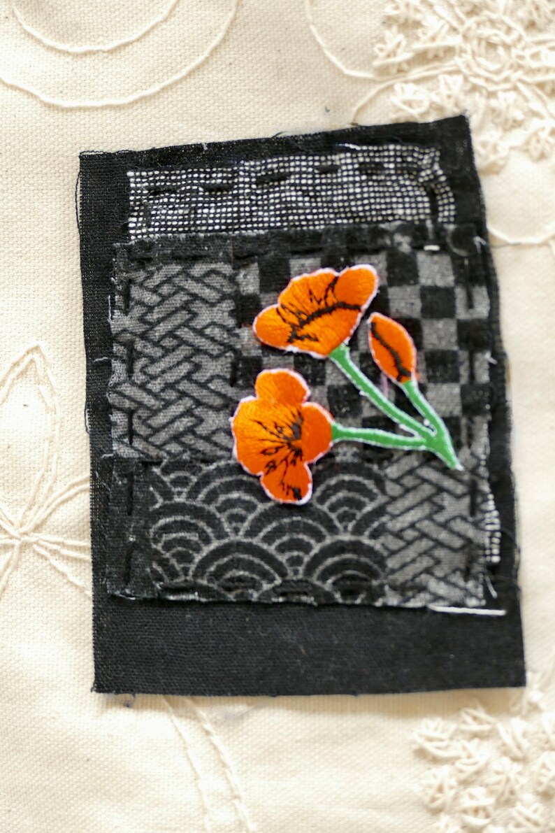 Peut inclure: Un carr&eacute; de tissu noir avec un motif patchwork pr&eacute;sentant diff&eacute;rents motifs. Une petite fleur orange brod&eacute;e avec des tiges vertes est centr&eacute;e sur le tissu.
