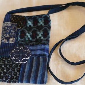 May include: A patchwork shoulder bag in various shades of blue. The bag features a mix of patterned fabrics, including stripes, floral, and geometric designs. A dark blue strap extends from the top of the bag. The bag is likely handmade.
