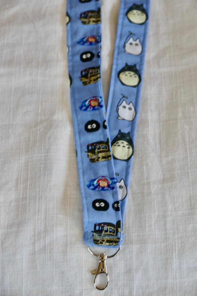 May include: A blue lanyard with a white clip. One side features a repeating pattern of Totoro and Catbus characters from the movie My Neighbor Totoro. The other side features a repeating pattern of No Face characters from the movie Spirited Away.