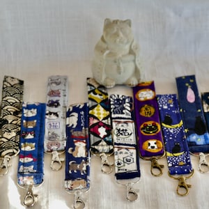 May include: A collection of 12 colorful keychains featuring various cat designs. The keychains are made of fabric and have a metal ring for attaching keys. The keychains are arranged in a row on a white surface.