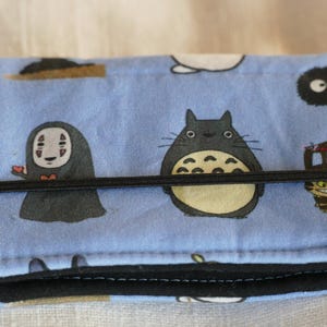 May include: A blue fabric wallet with a black elastic band closure. The wallet features a repeating pattern of cartoon characters from the animated film 'My Neighbor Totoro' and 'Spirited Away'.