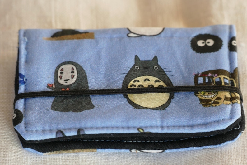 May include: A blue fabric wallet with a black elastic band closure. The wallet features a repeating pattern of cartoon characters from the animated film 'My Neighbor Totoro' and 'Spirited Away'.