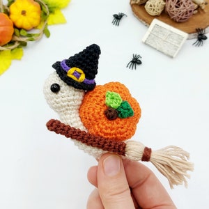 May include: A crocheted white snail wearing a black witch hat with a purple and yellow band, carrying a brown stick with a beige broom attached, and a crocheted orange pumpkin on its back.