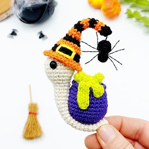 May include: A crocheted snail wearing a witch's hat with orange and black stripes, a purple shell, and a green scarf. The snail is holding a black spider with long legs.