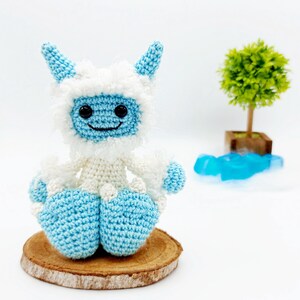 May include: A crocheted blue and white yeti sitting on a wooden slice. The yeti has a smiling face with black eyes and a white fluffy beard.