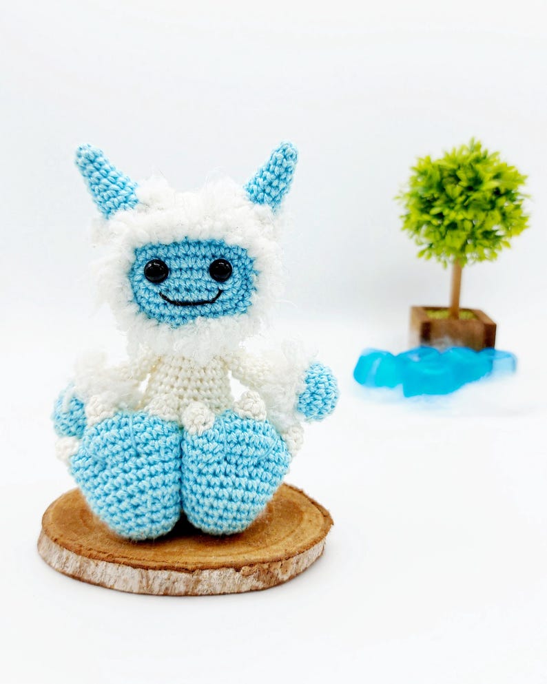 May include: A crocheted blue and white yeti sitting on a wooden slice. The yeti has a smiling face with black eyes and a white fluffy beard.