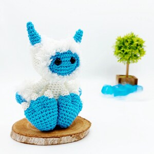 May include: A crocheted blue and white yeti toy sitting on a wooden slice. The yeti has a fluffy white body and blue horns, legs, and face.