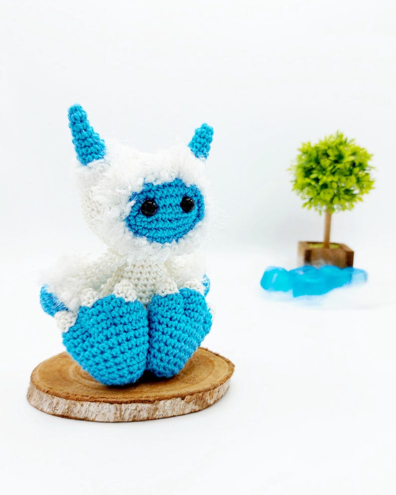 May include: A crocheted blue and white yeti toy sitting on a wooden slice. The yeti has a fluffy white body and blue horns, legs, and face.