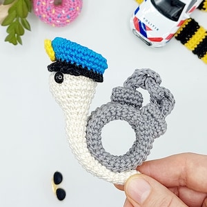 May include: Crocheted police-themed toys, including a white and gray snail wearing a blue and yellow police hat and gray handcuffs. A pink donut with sprinkles, toy police car, and sunglasses are also visible.