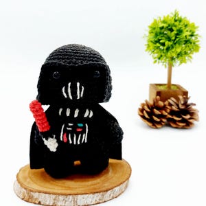Könnte beinhalten: Ein schwarzes, gehäkeltes Darth Vader Plüschtier, das ein rotes Lichtschwert hält. Das Spielzeug sitzt auf einer Holzscheibe.