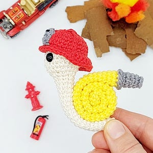 Puede incluir: Un caracol tejido a ganchillo con un gorro de bombero rojo y una manguera amarilla. También hay un camión de bomberos rojo en miniatura, una boca de incendios y un extintor. El caracol tiene un cuerpo blanco y detalles grises. El número 10 está en la cara del caracol.