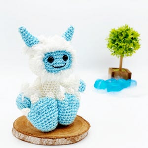 May include: A crocheted yeti toy with blue and white fur, sitting on a wooden slice. The yeti has a smiling face with black eyes and a white beard.