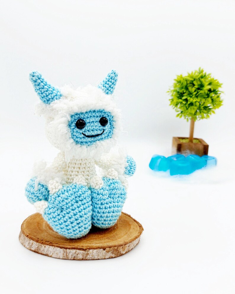 May include: A crocheted yeti toy with blue and white fur, sitting on a wooden slice. The yeti has a smiling face with black eyes and a white beard.