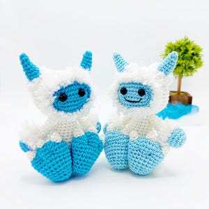 May include: Two crocheted yeti plush toys, one blue and one white, with black eyes and a smiling mouth. The yeti toys are sitting on a white surface.