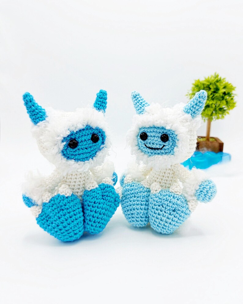 May include: Two crocheted yeti plush toys, one blue and one white, with black eyes and a smiling mouth. The yeti toys are sitting on a white surface.