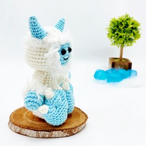 May include: A crocheted yeti toy with blue horns and feet, sitting on a wooden slice. The yeti is white with blue accents and has black eyes.