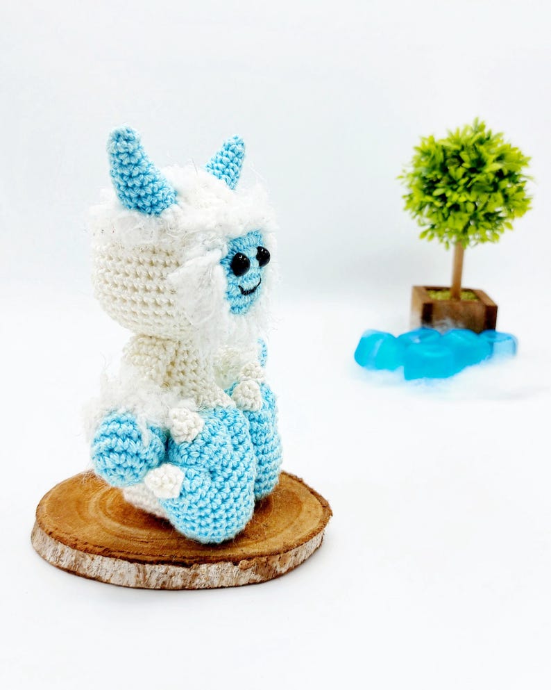 May include: A crocheted yeti toy with blue horns and feet, sitting on a wooden slice. The yeti is white with blue accents and has black eyes.