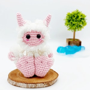 May include: A pink and white crocheted plush toy with a fluffy white body and pink horns, sitting on a wooden slice. The toy has black eyes and a pink face.