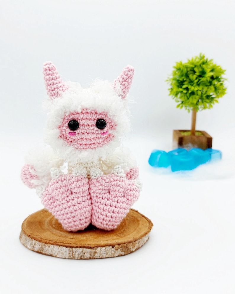May include: A pink and white crocheted plush toy with a fluffy white body and pink horns, sitting on a wooden slice. The toy has black eyes and a pink face.