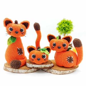 Op de afbeelding: Drie gehaakte pompoenkatten speelgoed, één zit rechtop, één zit met zijn staart om zijn lichaam gekruld en één zit met zijn hoofd naar voren gericht. De katten zijn oranje met bruine neuzen en zwarte ogen. Ze zitten op houten schijven.