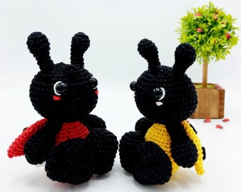 May include: Two handmade crochet ladybug toys. One ladybug has a red wing, the other has a yellow wing. Both are black with white eyes and small red cheeks. The toys are sitting on a white surface.