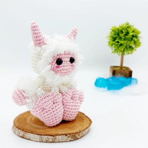 May include: A pink and white crocheted plush toy with horns, sitting on a wooden slice. The toy has black eyes and a pink nose.
