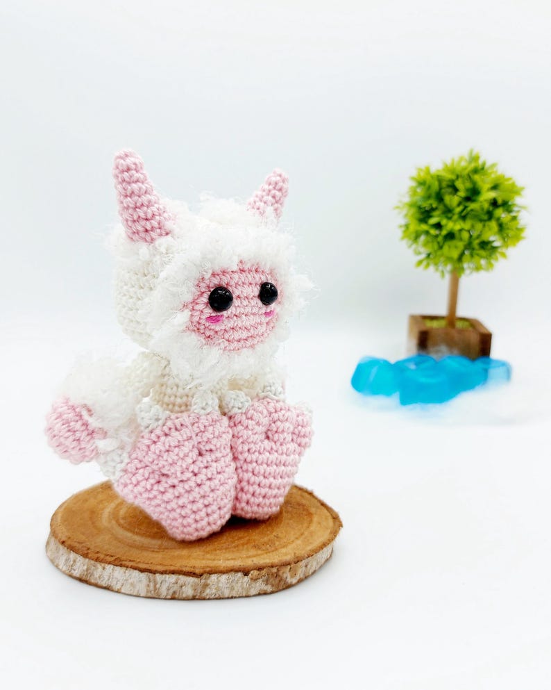 May include: A pink and white crocheted plush toy with horns, sitting on a wooden slice. The toy has black eyes and a pink nose.