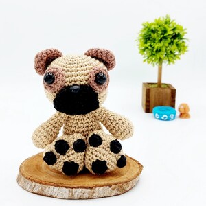 Op de afbeelding: Een handgemaakt gehaakt mopshondspeeltje in beige, bruin en zwart. De mopshond heeft grote zwarte ogen, een zwarte snuit en zwarte vlekken op zijn poten. Het staat op een klein houten schijfje. Een kleine groene plant in een houten pot staat op de achtergrond.