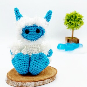 May include: A crocheted blue and white yeti toy with black eyes sitting on a wooden slice.