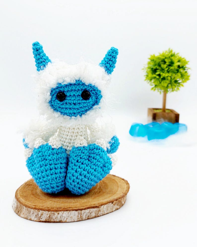 May include: A crocheted blue and white yeti toy with black eyes sitting on a wooden slice.