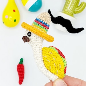 May include: A crocheted amigurumi toy shaped like a taco with a sombrero. The taco is yellow with green and red accents. The sombrero is yellow with red, green, blue, and yellow stripes. The toy has a white body and a black mustache.