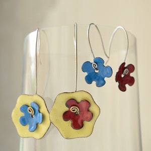 Peut inclure: Trois paires de boucles d'oreilles avec des breloques en forme de fleurs. Les breloques sont jaunes, bleues et rouges. Les boucles d'oreilles sont suspendues à une surface en verre.