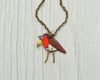 May include: A ceramic robin necklace with a brown body, red breast, and white accents. The robin is holding a small yellow bead in its beak. The necklace is made with a bronze chain.