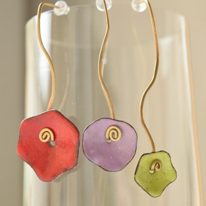 Peut inclure: Trois paires de boucles d'oreilles avec des pendentifs en forme de fleurs colorées et des accents de fil d'or. Les boucles d'oreilles sont suspendues à une surface en verre.