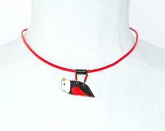 Könnte beinhalten: Eine Halskette aus rotem Band mit einem kleinen Vogel-Anhänger aus Emaille. Der Vogel ist schwarz, weiß und rot mit einem gelben Schnabel.