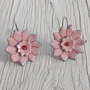 May include: A pair of handmade flower earrings. Each earring features a light pink, multi-petal flower design with red accents and a small red bead center. The earrings have silver-colored ear wires and are set against a white wooden background.