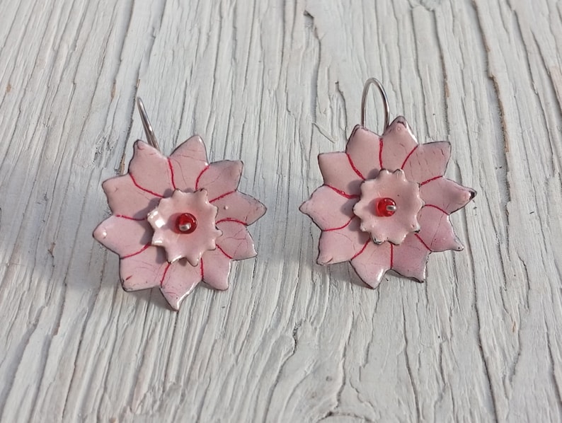 May include: A pair of handmade flower earrings. Each earring features a light pink, multi-petal flower design with red accents and a small red bead center. The earrings have silver-colored ear wires and are set against a white wooden background.