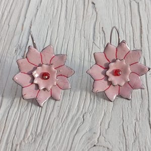 May include: A pair of handmade floral earrings. Each earring features a light pink, multi-petaled flower design with red accents and a small red bead in the center. The earrings have silver-colored hooks and are set against a white wooden surface.