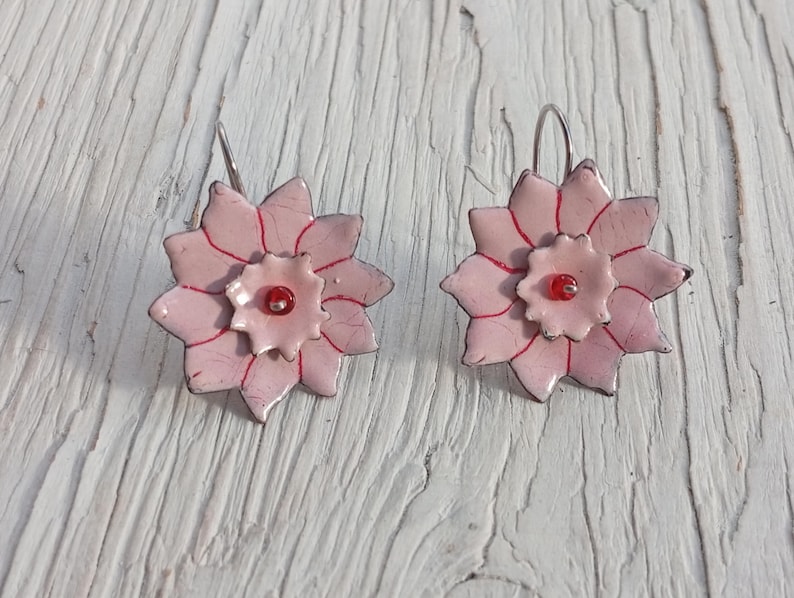 May include: A pair of handmade floral earrings. Each earring features a light pink, multi-petaled flower design with red accents and a small red bead in the center. The earrings have silver-colored hooks and are set against a white wooden surface.