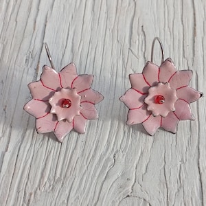 May include: A pair of handmade floral earrings. Each earring features a light pink, multi-petaled flower design with red accents and a small red bead center. The earrings have silver-colored ear wires.