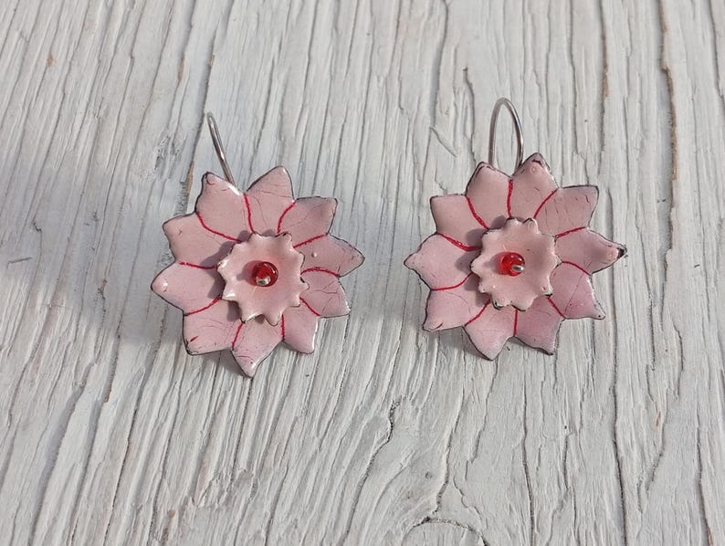 May include: A pair of handmade floral earrings. Each earring features a light pink, multi-petaled flower design with red accents and a small red bead center. The earrings have silver-colored ear wires.