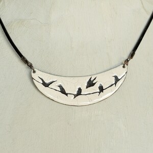 May include: A white ceramic necklace with a crescent moon shape pendant featuring a black silhouette of birds on a branch.
