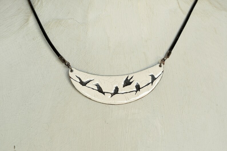 May include: A white ceramic necklace with a crescent moon shape pendant featuring a black silhouette of birds on a branch.