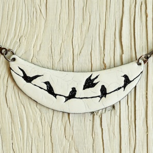May include: A white ceramic necklace with a crescent moon shape featuring black silhouettes of birds perched on a branch. The necklace is on a black leather cord.