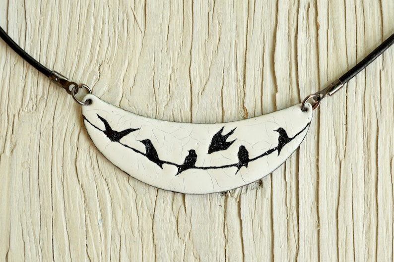 May include: A white ceramic necklace with a crescent moon shape featuring black silhouettes of birds perched on a branch. The necklace is on a black leather cord.