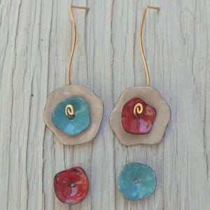 Peut inclure: Boucles d'oreilles artisanales avec un motif floral. Chaque boucle d'oreille a un fil doré et un design en couches avec une couche extérieure beige, un centre turquoise ou rouge et un accent en spirale doré. Sur une surface en bois clair.